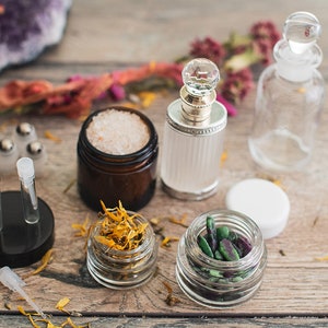 Op de afbeelding: Een verzameling kleine glazen potten en flessen met verschillende ingrediënten, waaronder gedroogde bloemen, zout, kristallen en kruiden. De potten zijn gerangschikt op een houten oppervlak met een paarse amethistkristal op de achtergrond.