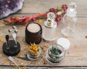 May include: A collection of small glass jars and bottles containing various ingredients, including dried flowers, salt, crystals, and herbs. The jars are arranged on a wooden surface with a purple amethyst crystal in the background.