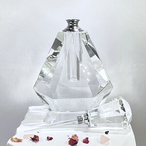 May include: A clear glass bottle with a faceted design and a silver top. The bottle is empty and has a clear glass dropper attached to it. The bottle is sitting on a white surface with some pink rose petals.