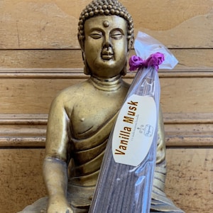 May include: A package of brown incense sticks with a white label that reads "Vanilla Musk" with natural incense inside. The incense is sitting in front of a golden statue of Buddha.