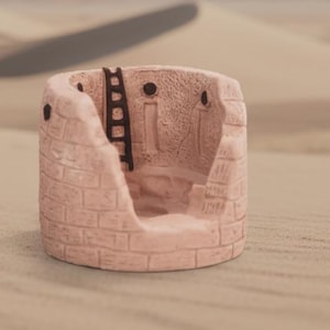 May include: A small, pink, cylindrical decorative planter with a weathered brick design. The planter has a partial wall and a black ladder. The background is a blurred desert scene.