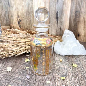 May include: A clear glass bottle with a gold and white floral design. The bottle has a glass stopper and is decorated with gold leaf.