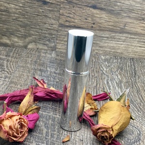 A silver metallic 12 ml spray atomizer bottle with roses surrounding it on wood. Elegant perfume travel bottle. The whole body is silver. This is an empty refillable bottle. Keep refilling it with your favorite perfume oil!