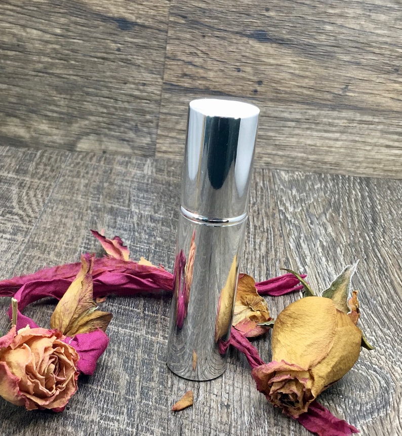 A silver metallic 12 ml spray atomizer bottle with roses surrounding it on wood. Elegant perfume travel bottle. The whole body is silver. This is an empty refillable bottle. Keep refilling it with your favorite perfume oil!