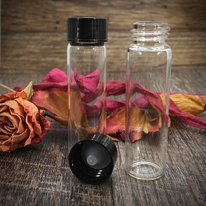 Op de afbeelding: Twee heldere glazen flessen met zwarte deksels. De flessen zijn leeg en hebben een cilindrische vorm. De flessen staan op een houten oppervlak.