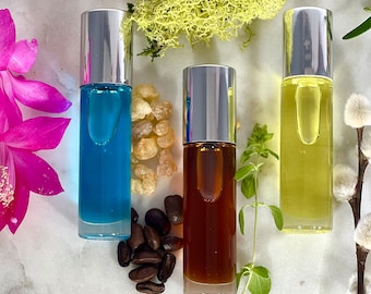 May include: Three glass bottles with silver caps filled with different colored liquids. The bottles are on a white marble surface with coffee beans, green foliage, and a pink flower.