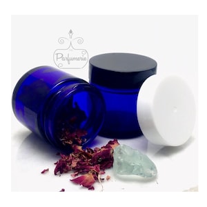 Pode incluir: Dois frascos de vidro azul cobalto com tampas pretas e uma tampa branca. Os frascos estão vazios e estão sobre uma superfície branca. Há pétalas de rosa secas e um cristal verde claro na superfície.
