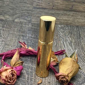 A gold metallic 12 ml spray atomizer bottle with roses surrounding it on wood. Elegant perfume travel bottle. The whole body is gold. This is an empty refillable bottle. Keep refilling it with your favorite perfume oil!