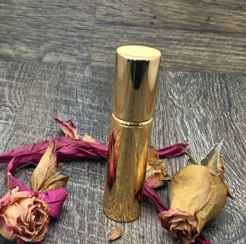 A gold metallic 12 ml spray atomizer bottle with roses surrounding it on wood. Elegant perfume travel bottle. The whole body is gold. This is an empty refillable bottle. Keep refilling it with your favorite perfume oil!