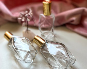 May include: Three clear glass bottles with gold tops. The bottles are faceted and have a geometric shape. The bottles are on a white surface with a pink crystal.
