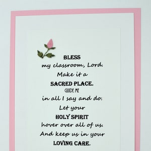 May include: A white card with a pink border and a small pink flower in the top left corner. The card has a poem printed on it that reads: "BLESS my classroom, Lord. Make it a SACRED PLACE. GUIDE ME in all I say and do. Let your HOLY SPIRIT hover over all of us. And keep us in your LOVING CARE."