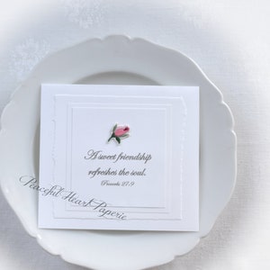 May include: A white card with a pink rose illustration and the text "A sweet friendship refreshes the soul. Proverbs 27:9" sits on a white plate. The card has a layered design with a torn edge. The words "Peaceful Heart Paperie" are also visible.