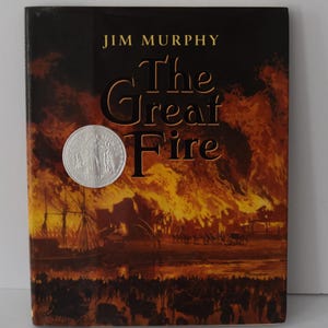 May include: A hardcover book titled "The Great Fire" by Jim Murphy. The cover depicts a fiery scene with a silver circular sticker. The book's cover is predominantly dark with orange and brown hues, suggesting a historical or disaster theme.