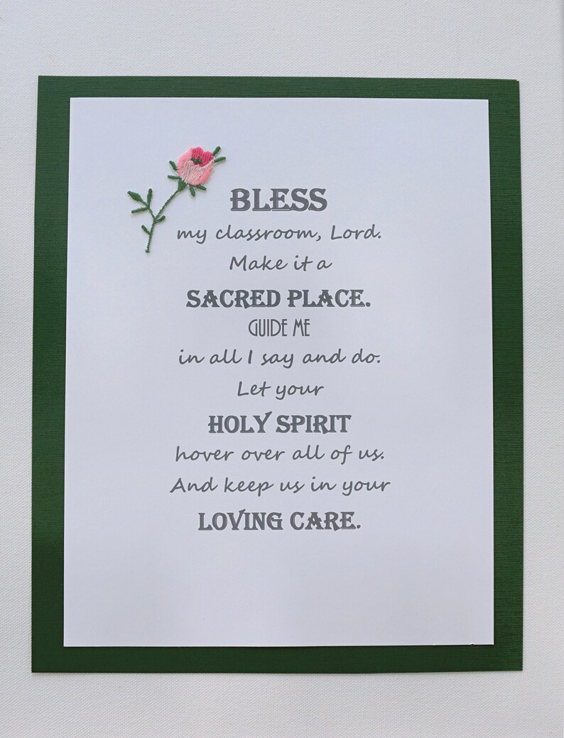 May include: A white card with a green border and a small pink flower embroidered in the top right corner. The card has a poem printed on it that reads: "BLESS my classroom, Lord. Make it a SACRED PLACE. GUIDE ME in all I say and do. Let your HOLY SPIRIT hover over all of us. And keep us in your LOVING CARE."