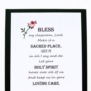 May include: A framed print with a pink flower embroidered in the top left corner. The print features a poem that reads: "BLESS my classroom, Lord. Make it a SACRED PLACE. GUIDE ME in all I say and do. Let your HOLY SPIRIT hover over all of us. And keep us in your LOVING CARE."