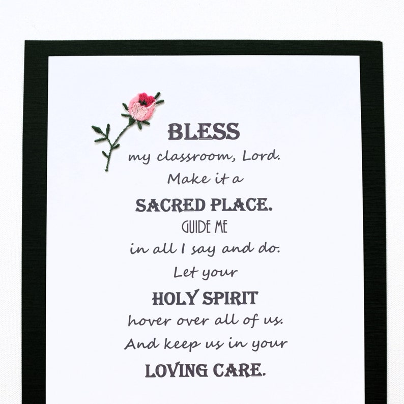 May include: A framed print with a pink flower embroidered in the top left corner. The print features a poem that reads: "BLESS my classroom, Lord. Make it a SACRED PLACE. GUIDE ME in all I say and do. Let your HOLY SPIRIT hover over all of us. And keep us in your LOVING CARE."
