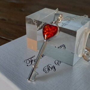 Puede incluir: Un collar de plata con un colgante en forma de corazón rojo. El colgante está hecho de ópalo y tiene un acabado brillante. El collar se muestra en una caja blanca con las palabras "Forever By Q" escritas en cursiva negra.