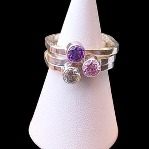 May include: Three silver rings with colourful gemstones. The rings are stacked on a white cone-shaped ring holder. The gemstones are purple, pink, and green.