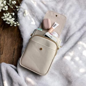 May include: A beige crossbody bag with a gold zipper and button closure, open to reveal a phone, makeup, and a pair of wireless earbuds. The bag is on a soft, gray blanket, with white flowers in the background.