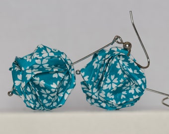 May include: A pair of earrings featuring teal fabric flowers with white floral patterns. The fabric flowers are attached to silver-colored metal wires. The earrings are displayed against a white background.