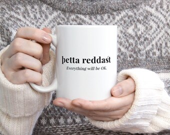 Könnte beinhalten: Weiße Keramiktasse mit schwarzem Text, der "betta reddast" und "Alles wird gut." lautet.