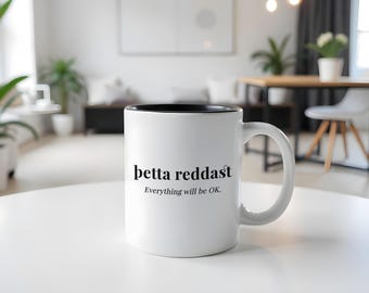 Könnte beinhalten: Weiße Keramik-Kaffeetasse mit schwarzem Rand. Die Tasse trägt den Text "betta reddast" und "Everything will be OK."