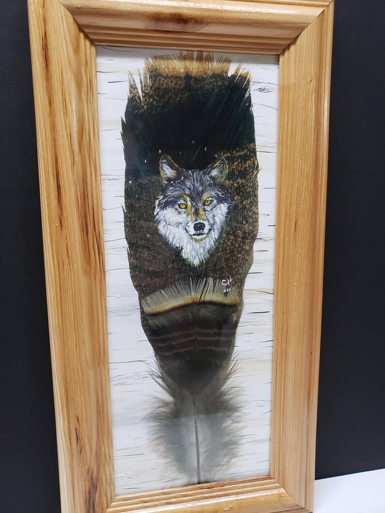 以下が含まれることがあります： 大きな羽に描かれたオオカミが特徴の額装されたアートワーク。オオカミは灰色の毛皮と黄色の目をしています。羽には茶色、黒、白の模様があります。フレームは明るい色の木材で作られています。