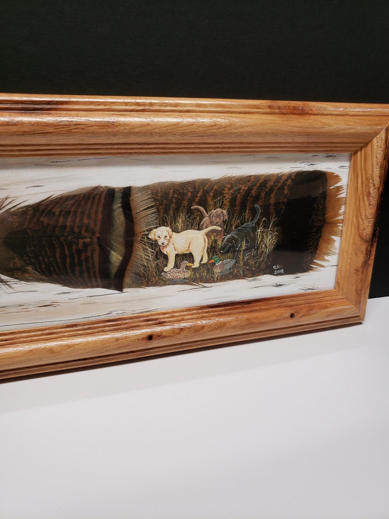 May include: A framed painting of a turkey feather with a scene of four Labrador Retriever puppies painted inside. The puppies are brown and yellow and are surrounded by green grass and a few small birds. The painting is signed "C.P. 2018".