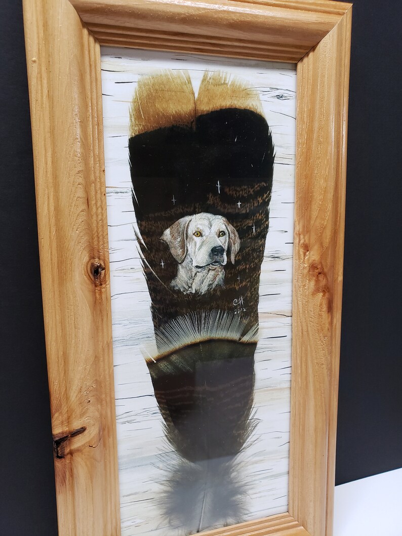May include: A framed painting of a white dog with brown spots, looking directly at the viewer. The dog is painted on a brown and black feather with a white background. The painting is signed "C.H. 2018".
