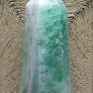 May include: A green and white fluorite crystal point, also known as a wand, with a smooth, polished surface. The crystal is a natural formation with a unique pattern of green and white.