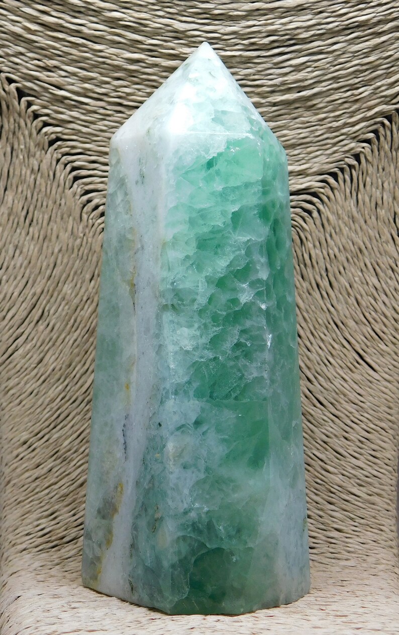 May include: A green and white fluorite crystal point, also known as a wand, with a smooth, polished surface. The crystal is a natural formation with a unique pattern of green and white.