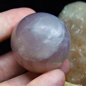 May include: A smooth, polished amethyst sphere with a light purple hue and white inclusions. The sphere is being held in a hand.