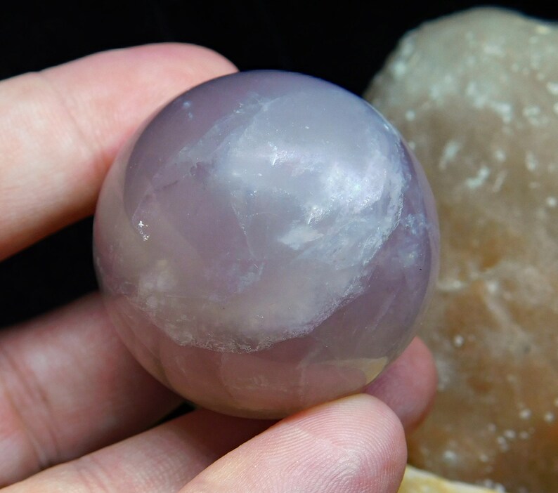 May include: A smooth, polished amethyst sphere with a light purple hue and white inclusions. The sphere is being held in a hand.