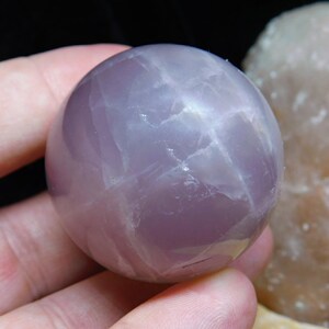 May include: A smooth, polished, lavender amethyst sphere with a light purple hue and white streaks. The sphere is approximately 2 inches in diameter.