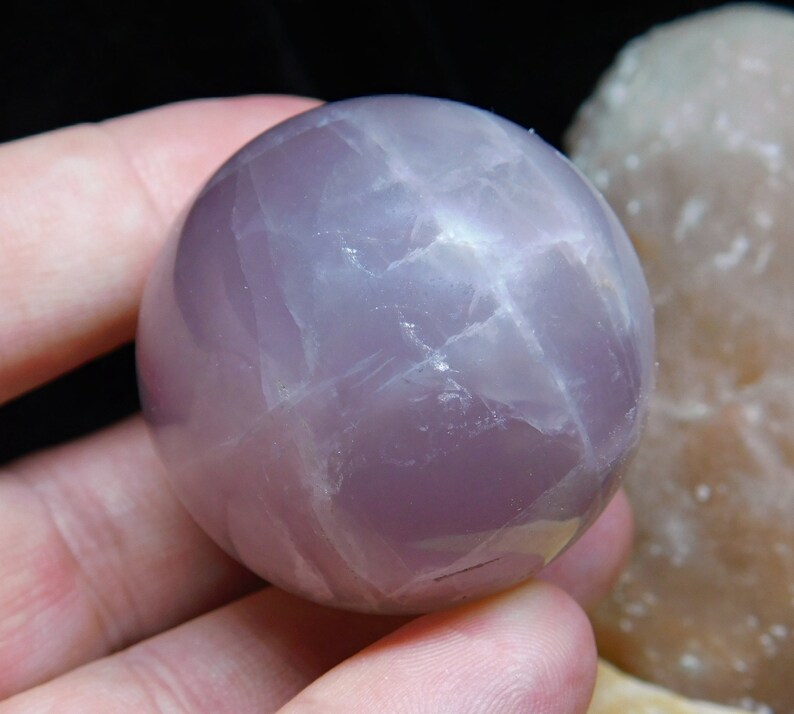 May include: A smooth, polished, lavender amethyst sphere with a light purple hue and white streaks. The sphere is approximately 2 inches in diameter.