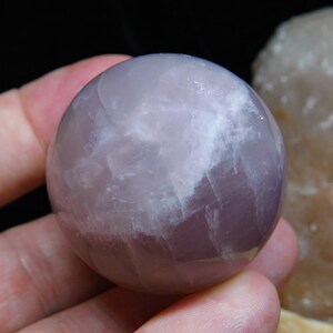 May include: A polished amethyst sphere with a light purple and white color pattern. The sphere is smooth and has a glossy finish.
