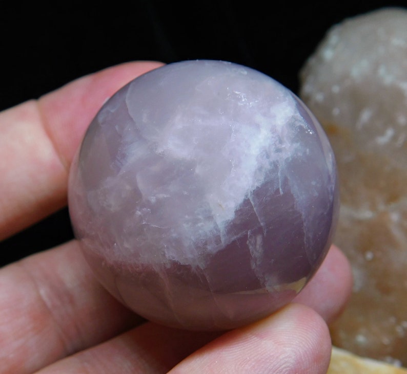 May include: A polished amethyst sphere with a light purple and white color pattern. The sphere is smooth and has a glossy finish.