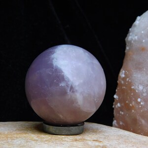 May include: A large, smooth, lavender amethyst sphere on a black stand. The sphere has a white, cloudy appearance.
