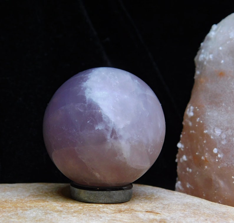 May include: A large, smooth, lavender amethyst sphere on a black stand. The sphere has a white, cloudy appearance.