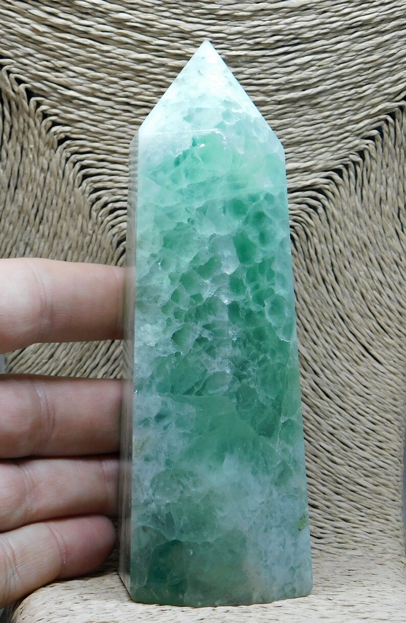 May include: A green fluorite crystal point, also known as a wand, with a light green and white color pattern. The crystal is polished and has a smooth, glassy surface.