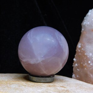 May include: A large, polished, pale pink rose quartz sphere on a black stand. The sphere is translucent and has a cloudy, swirling pattern.