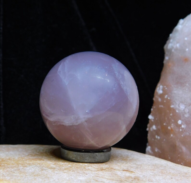 May include: A large, polished, pale pink rose quartz sphere on a black stand. The sphere is translucent and has a cloudy, swirling pattern.