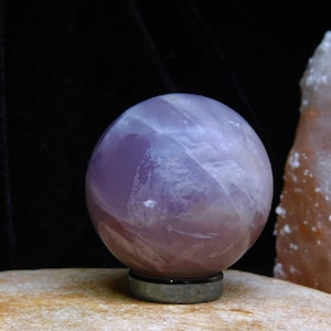 May include: A polished amethyst sphere with a swirling purple and white pattern. The sphere is sitting on a black base.