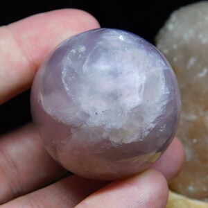 May include: A smooth, polished, lavender fluorite sphere with a white, cloudy interior. The sphere is approximately 2 inches in diameter.