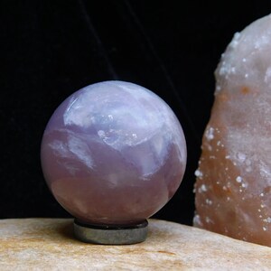 May include: A purple amethyst sphere with white swirls and a polished finish. The sphere is sitting on a black base.