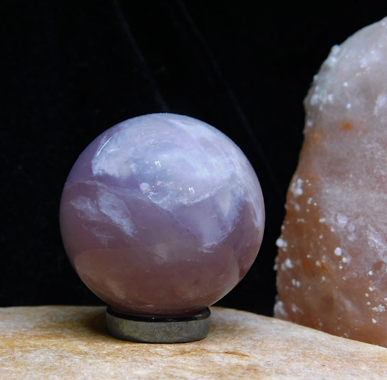 May include: A purple amethyst sphere with white swirls and a polished finish. The sphere is sitting on a black base.