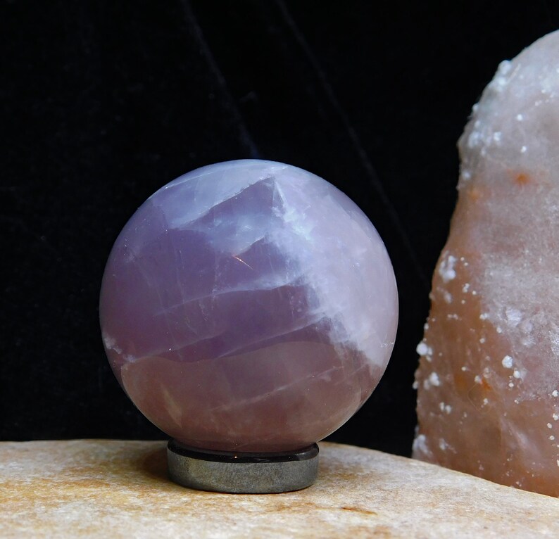 May include: A smooth, lavender-colored crystal sphere on a black stand. The sphere has a translucent, cloudy appearance.