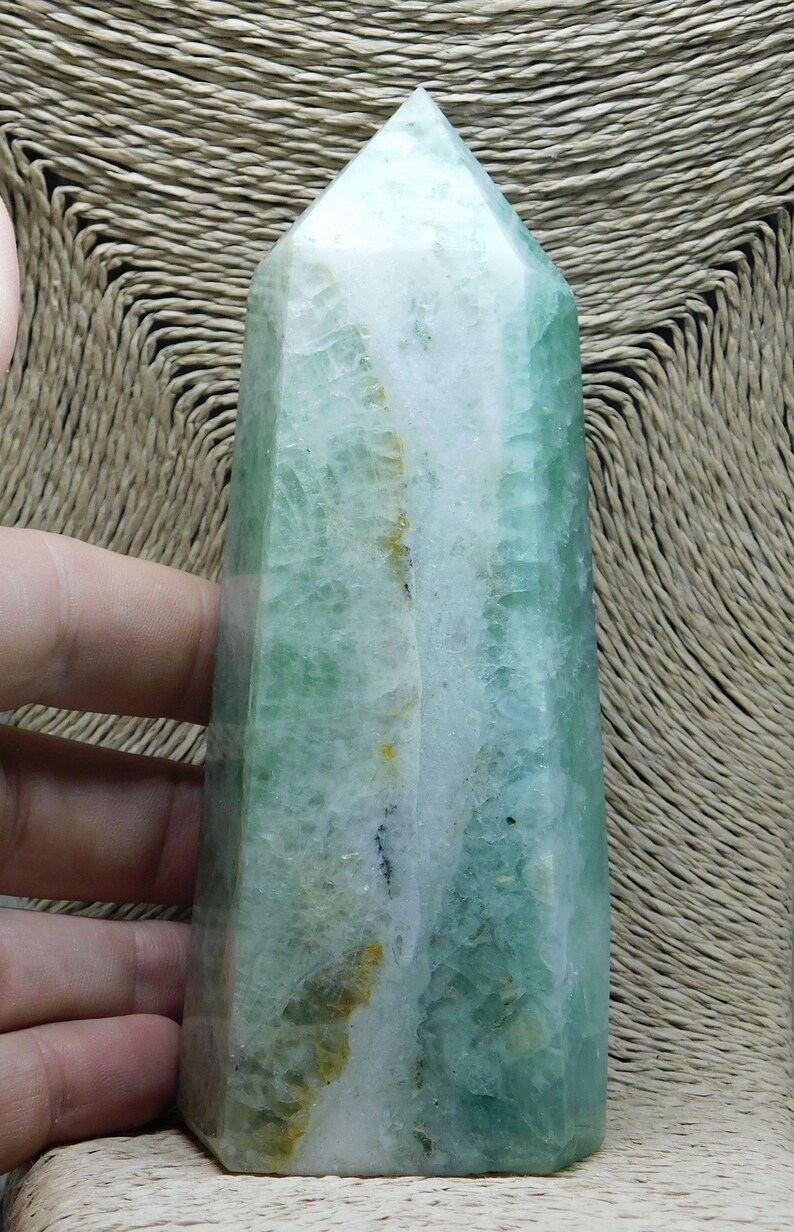May include: A green and white fluorite crystal point, also known as a wand, with a natural, raw finish. The crystal is translucent and has a striated pattern.