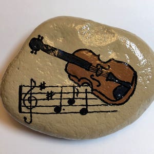 Puede incluir: Una piedra pintada a mano con un viol&iacute;n marr&oacute;n y notas musicales sobre un fondo beige.
