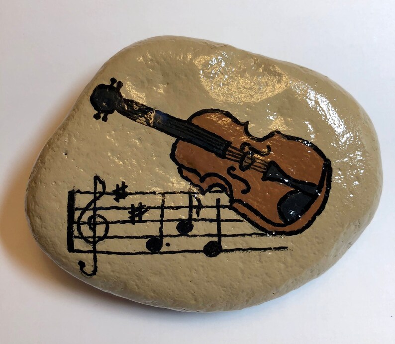 Puede incluir: Una piedra pintada a mano con un viol&iacute;n marr&oacute;n y notas musicales sobre un fondo beige.